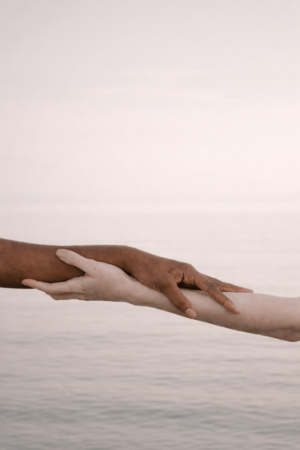 Two hands of different skin tones gently holding each other over the ocean at sunset, symbolizing connection, inclusion, and empathy