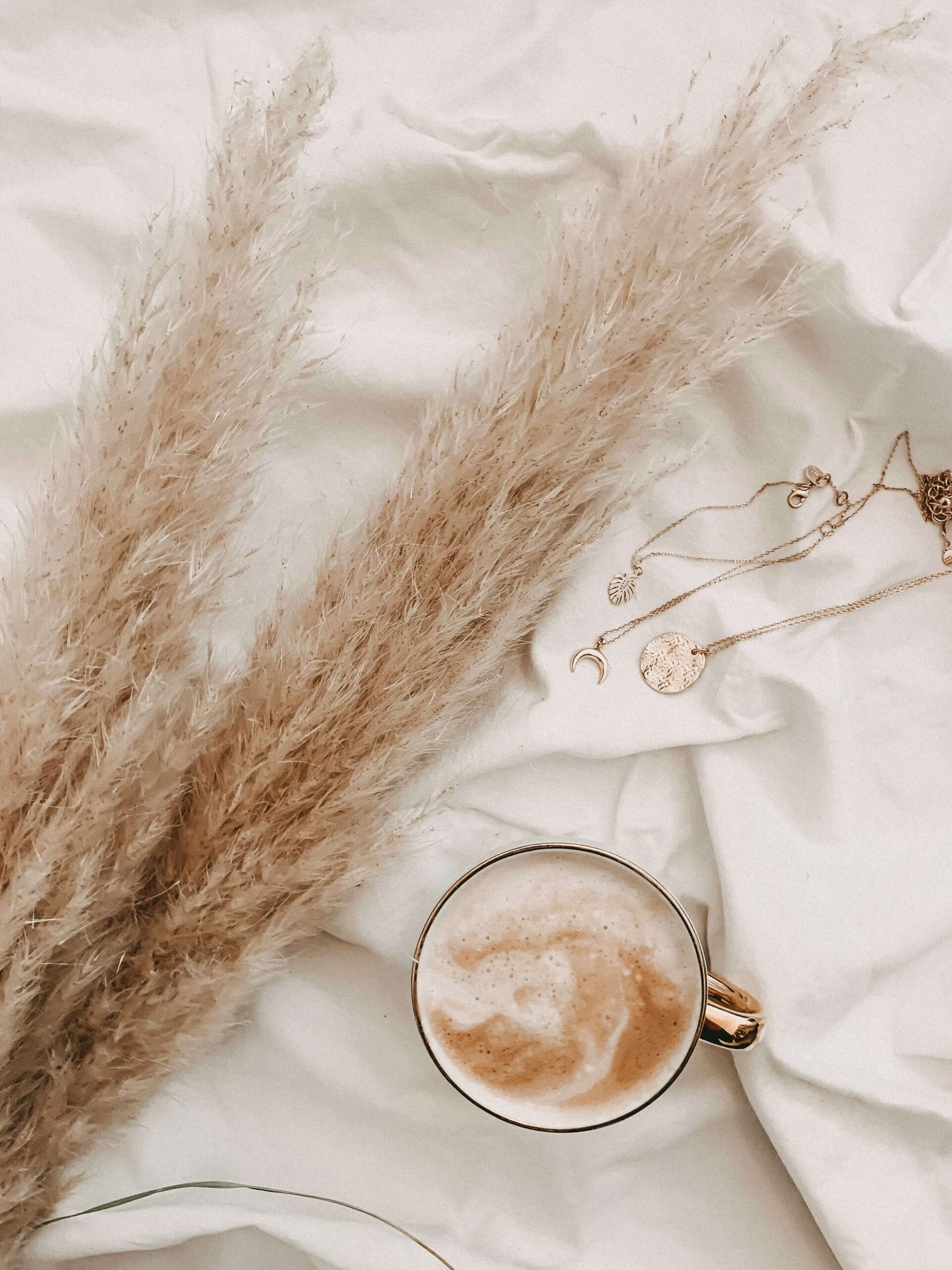 Latte, pampas grass, and gold jewelry on neutral fabric representing a calm writing environment for About page prompts